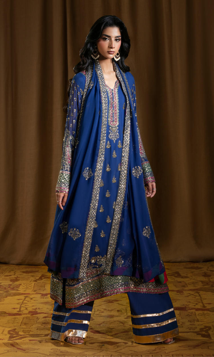 A woman modeling a royal blue traditional South Asian outfit with intricate gold and silver embroidery, paired with matching wide-leg pants and a draped dupatta, set against a warm brown draped backdrop.